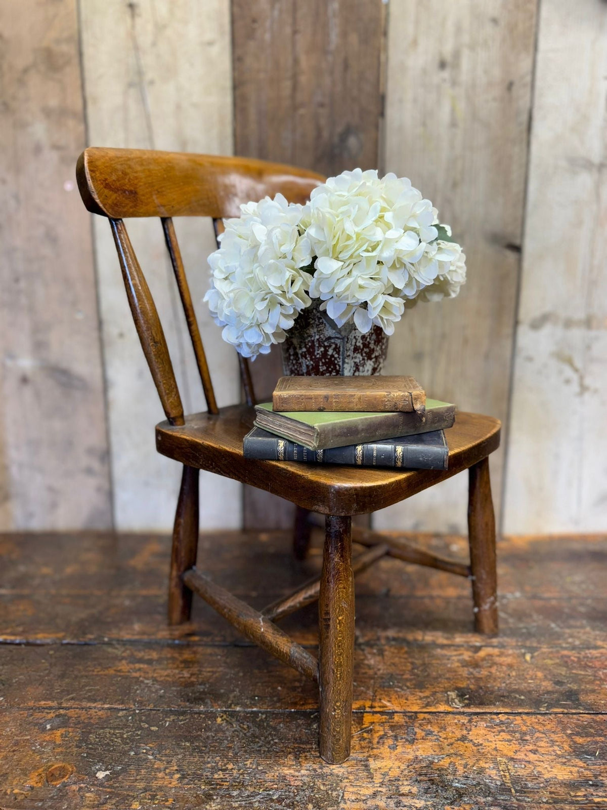 Antique childs chair with elm seat in rustic wood holding vintage books and white hydrangeas for nursery display