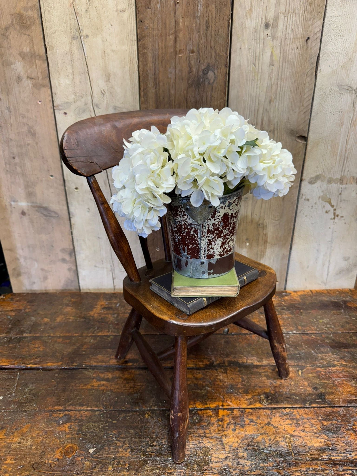 A rustic galvanized metal bucket with white hydrangeas next to an antique child’s chair with oak seat and distressed finish