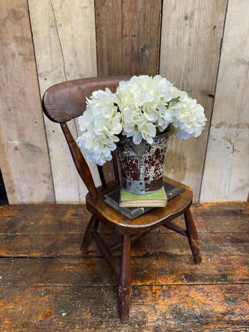 A rustic galvanized metal bucket with white hydrangeas next to an antique child’s chair with oak seat and distressed finish
