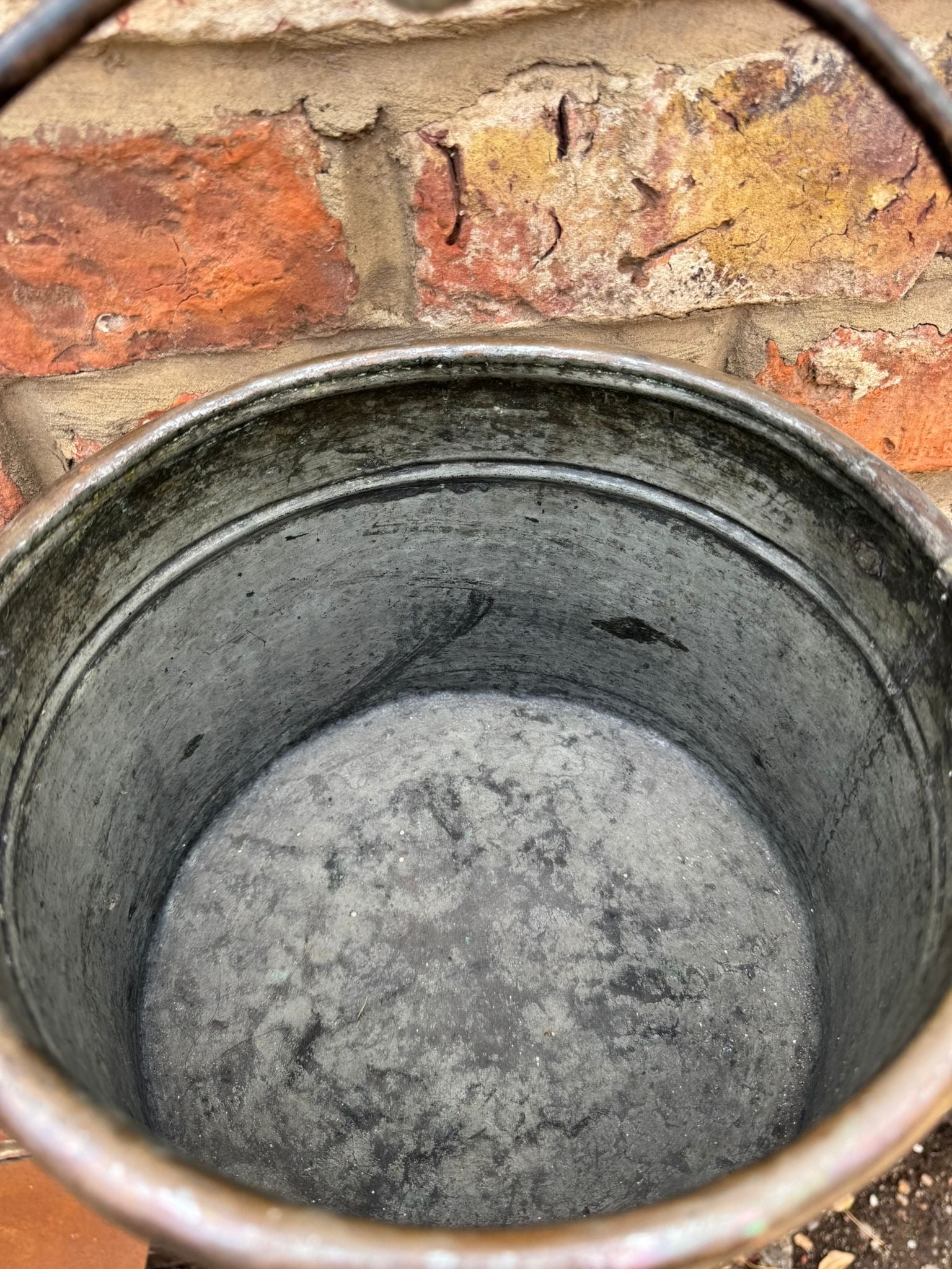Weathered black metal bucket with handle resting against brick wall resembling an antique copper cauldron pot