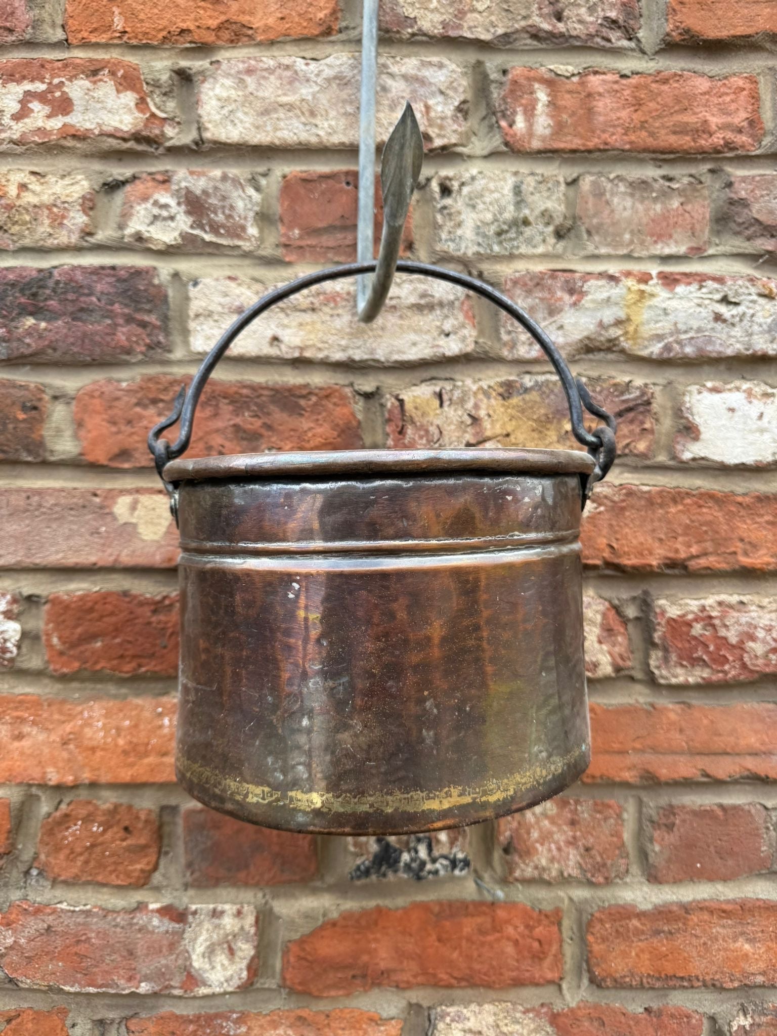 Antique copper cauldron pot with dark patina and black wrought iron handle hanging against brick wall