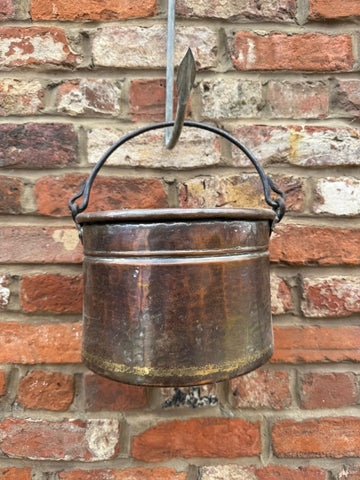 Antique copper cauldron pot with dark patina and black wrought iron handle hanging against brick wall