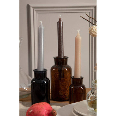 Three tall candles in gray brown and beige stand in dark amber black glass reagent bottles for apothecary use