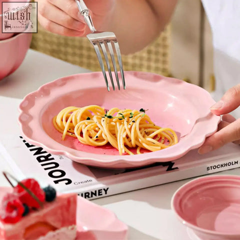 A pink ceramic plate with scalloped edges holding herb garnished spaghetti from the aria dining set
