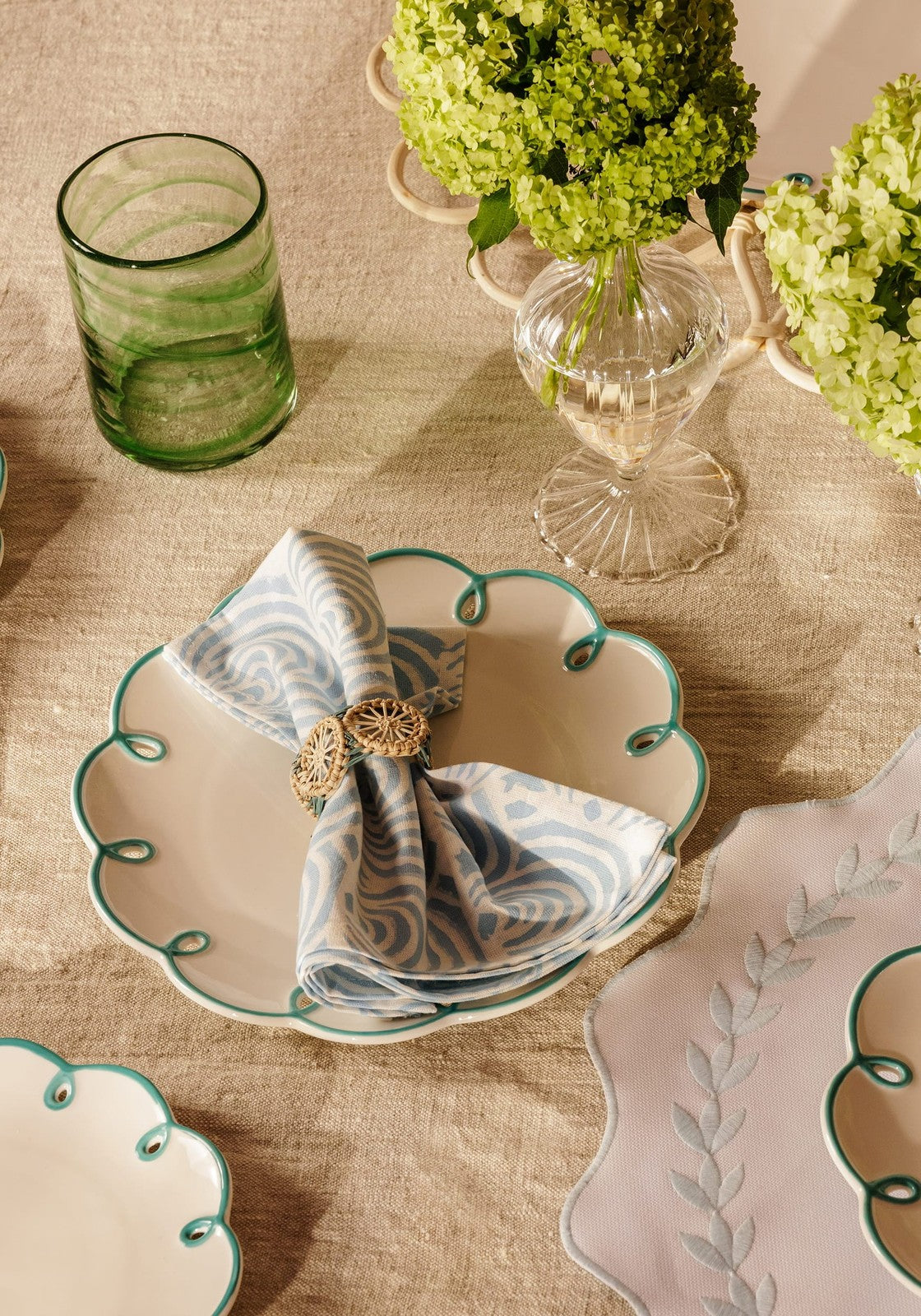 A delicate white ceramic plate with scalloped edge holds a blue and white atlántico iraca woven napkin ring in gold