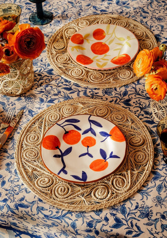 White ceramic plates with hand-painted orange fruit and leaves in vibrant colors resting on natural diamond weave rattan placemats