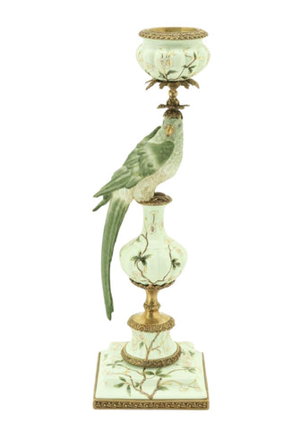 A delicate green parrot figurine perched on an ornate white porcelain taper candleholder with gold accents and floral patterns
