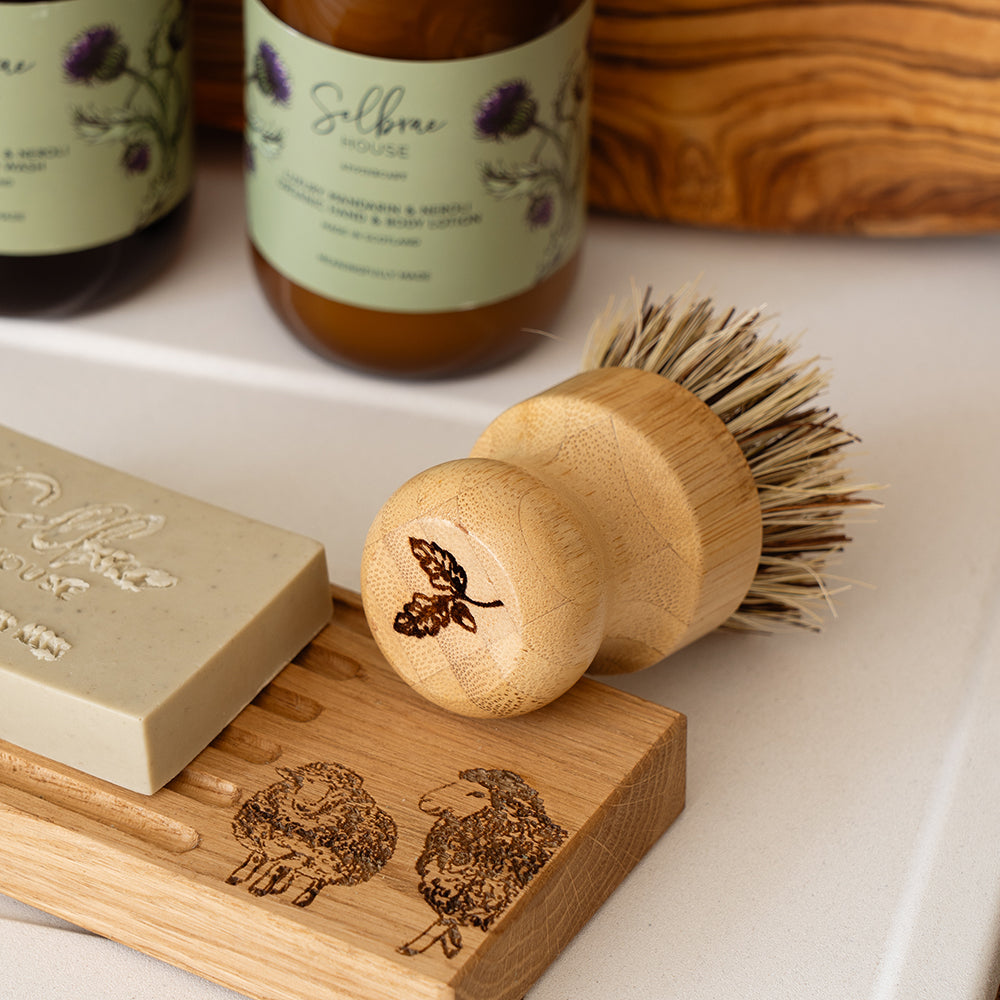 A light wood soap dish with engraved sheep and a matching bamboo pot brush with carved leaf handle design