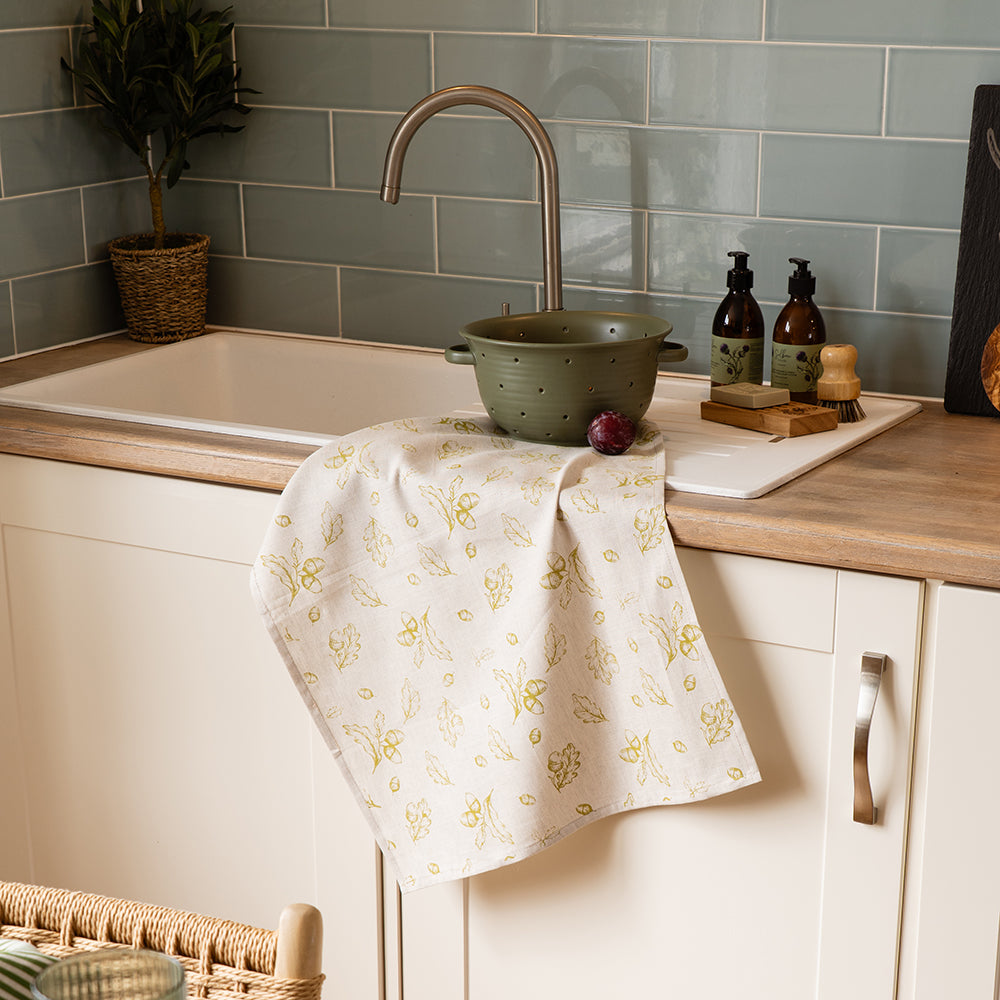 A cream-colored linen tea towel with a delicate yellow autumn acorns print hangs over a wooden countertop edge