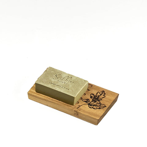A rectangular olive green oak soap resting on a light wood oak soap dish with engraved leaf and acorn design