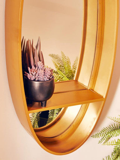 Avento gold oval wall mirror with gold metallic frame and built-in shelf holding a black ceramic pot with succulents