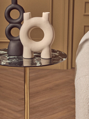 Cream-colored ceramic vase with central ring and three spouts on a side table with black marble effect top and gold base