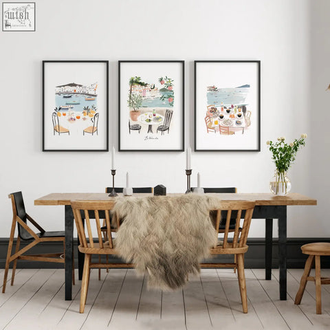 Three framed beach art prints of coastal mediterranean dining scenes hang above a wooden dining room table wall art set