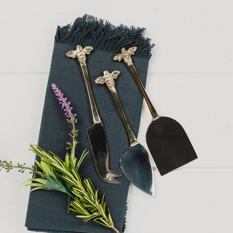 Set of three gold-toned bee cheese knives with black handles resting on a dark green napkin for cheese board gifts