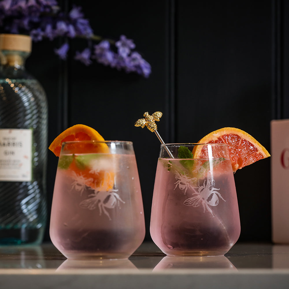 Pink etched bee-themed stemless glasses filled with pink cocktail and citrus slices with bee drinks stirrer