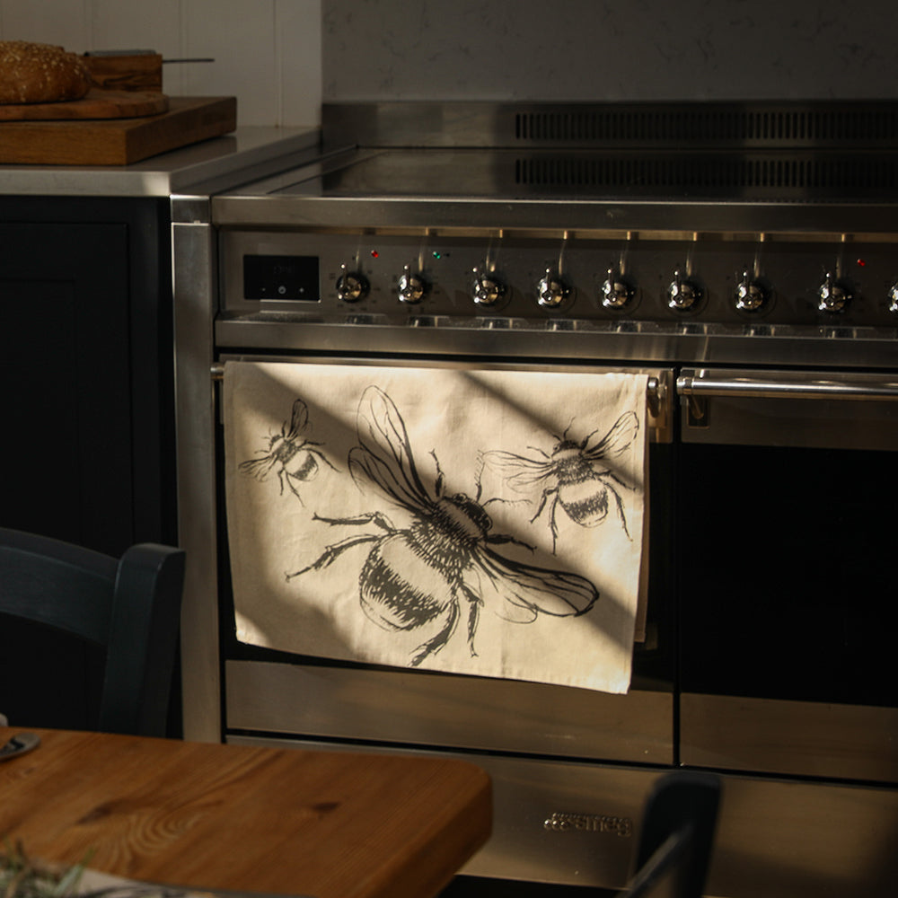 Beige linen tea towel with black bee illustrations hanging from a stainless steel oven handle for kitchen use