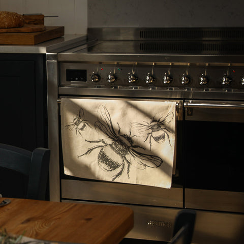 Beige linen tea towel with black bee illustrations hanging from a stainless steel oven handle for kitchen use