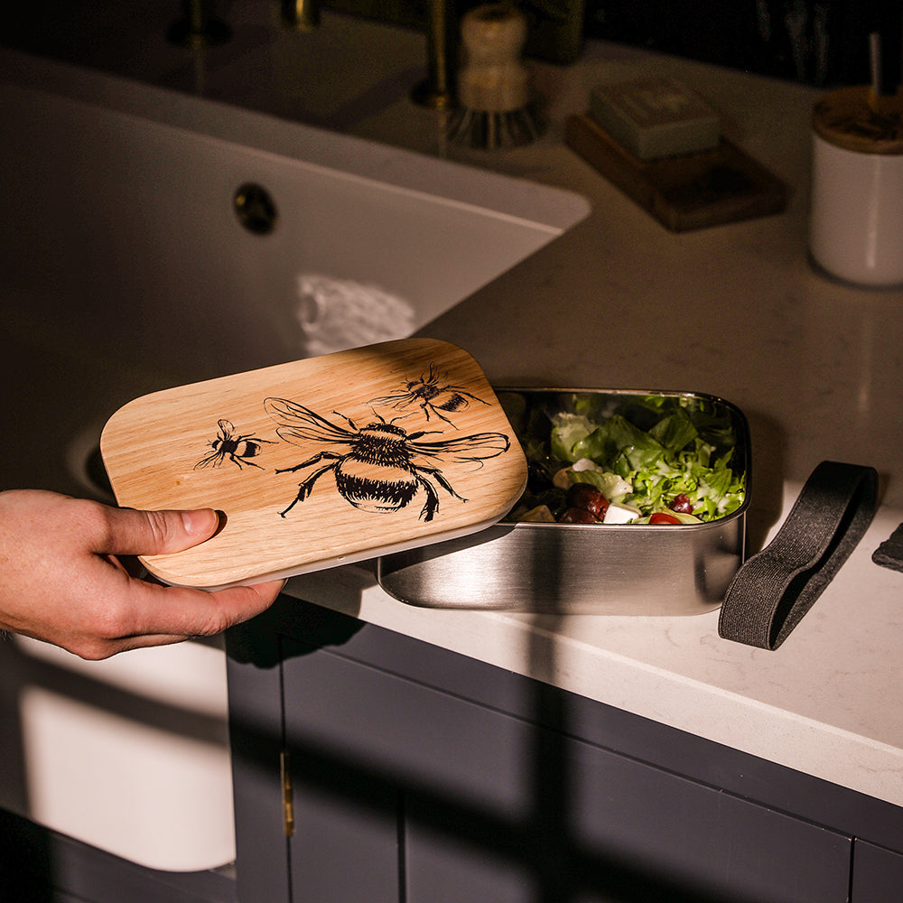 A stainless steel lunch box with a light wood lid engraved with bees filled with fresh salad greens