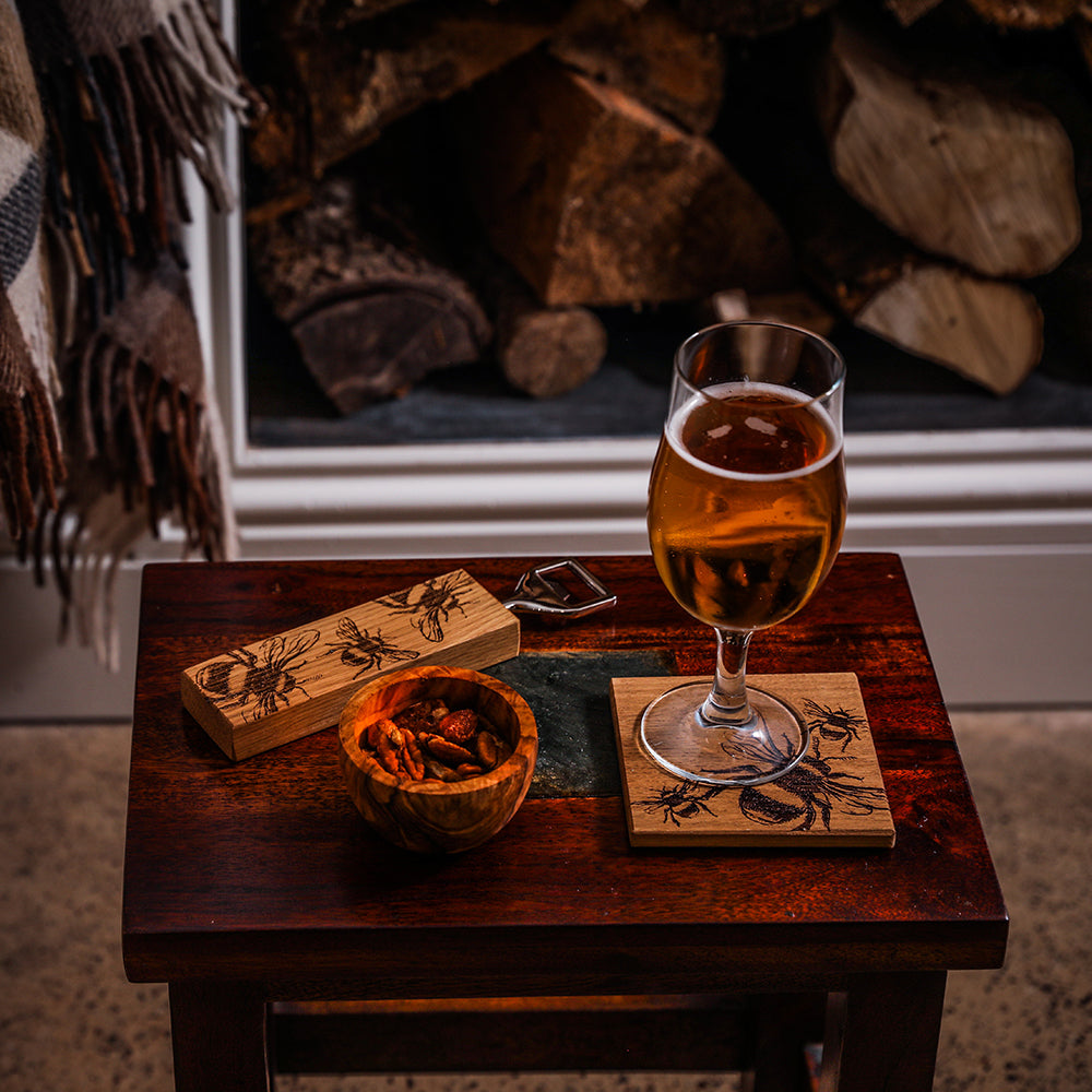 A rustic dark brown british oak coaster with an engraved bee design under a glass of amber beer from bee oak coasters set
