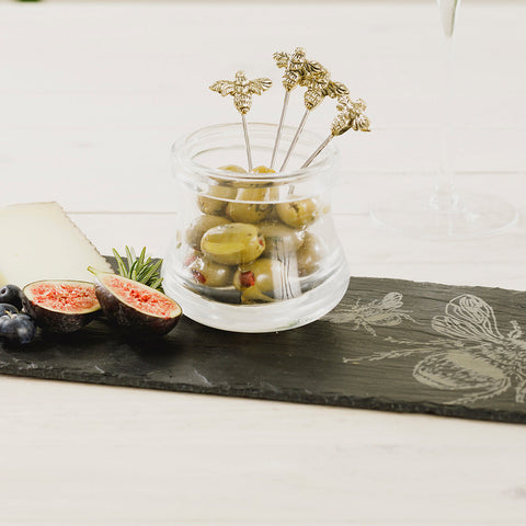 Clear glass jar filled with green olives topped with golden bee olive picks perfect for serving snacks