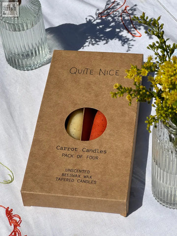 A brown cardboard box labeled quite nice containing a pack of four unscented cream and orange beeswax tapered candles