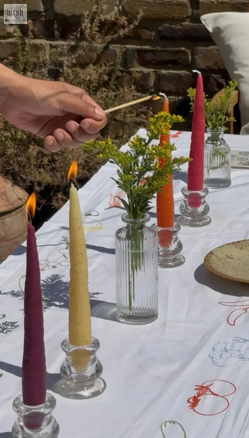 A hand holds a lit match near vibrant deep purple and golden yellow beeswax rainbow carrot stick candles in clear ribbed glass holders