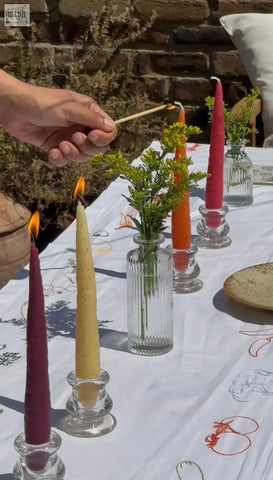 A hand holds a lit match near vibrant deep purple and golden yellow beeswax rainbow carrot stick candles in clear ribbed glass holders