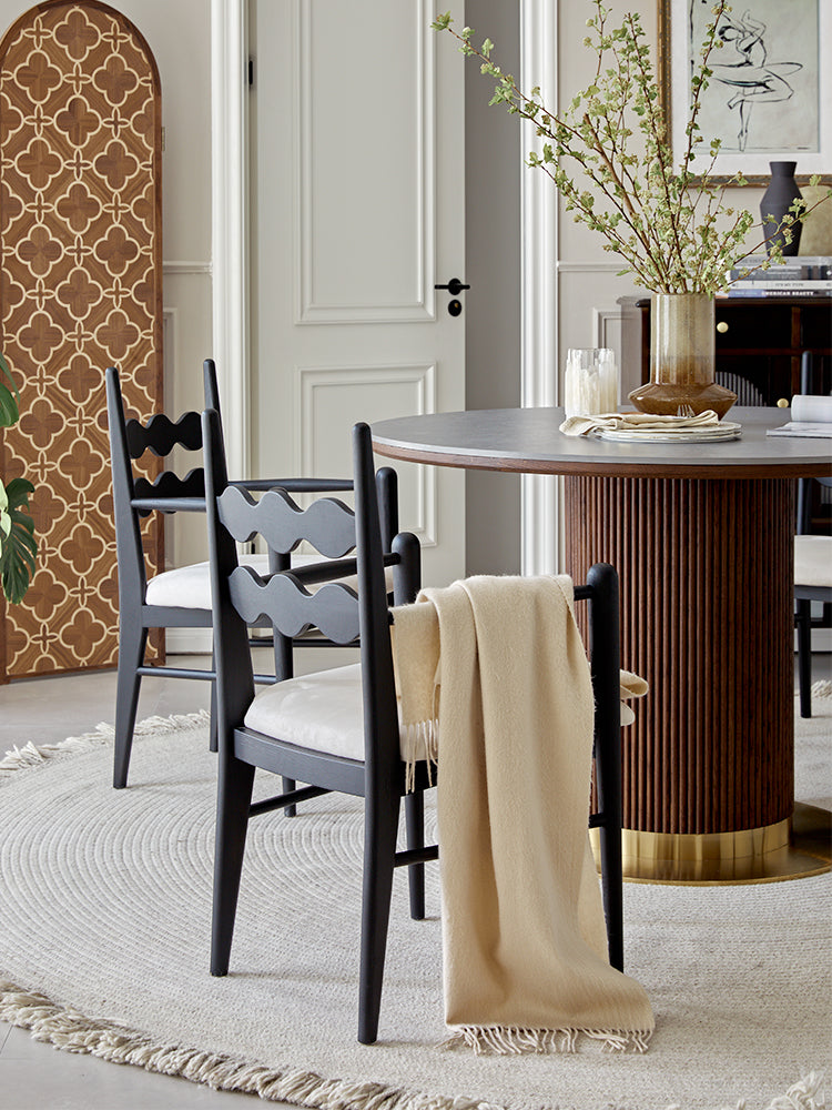 Dark gray wooden dining chair with wavy backrest and white cushion next to bek round dining table brown with beige throw