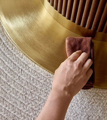 A hand wipes the curved brushed brass base of the bek round dining table brown with a soft terracotta cloth