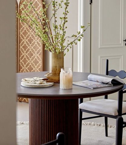 A gold-toned vase with green branches on a brown bek round dining table perfect for round dining spaces