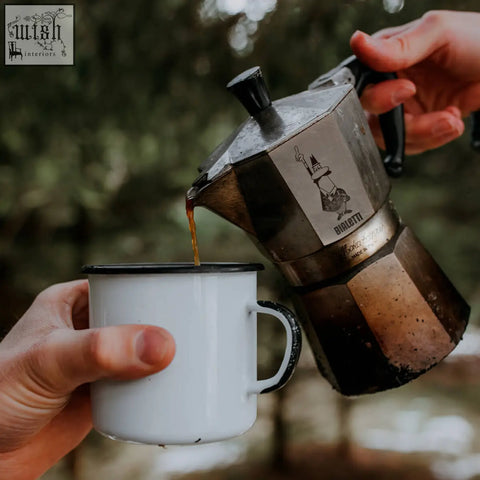 A vintage-style metallic bialetti moka express pouring espresso into a white enamel mug with black accents