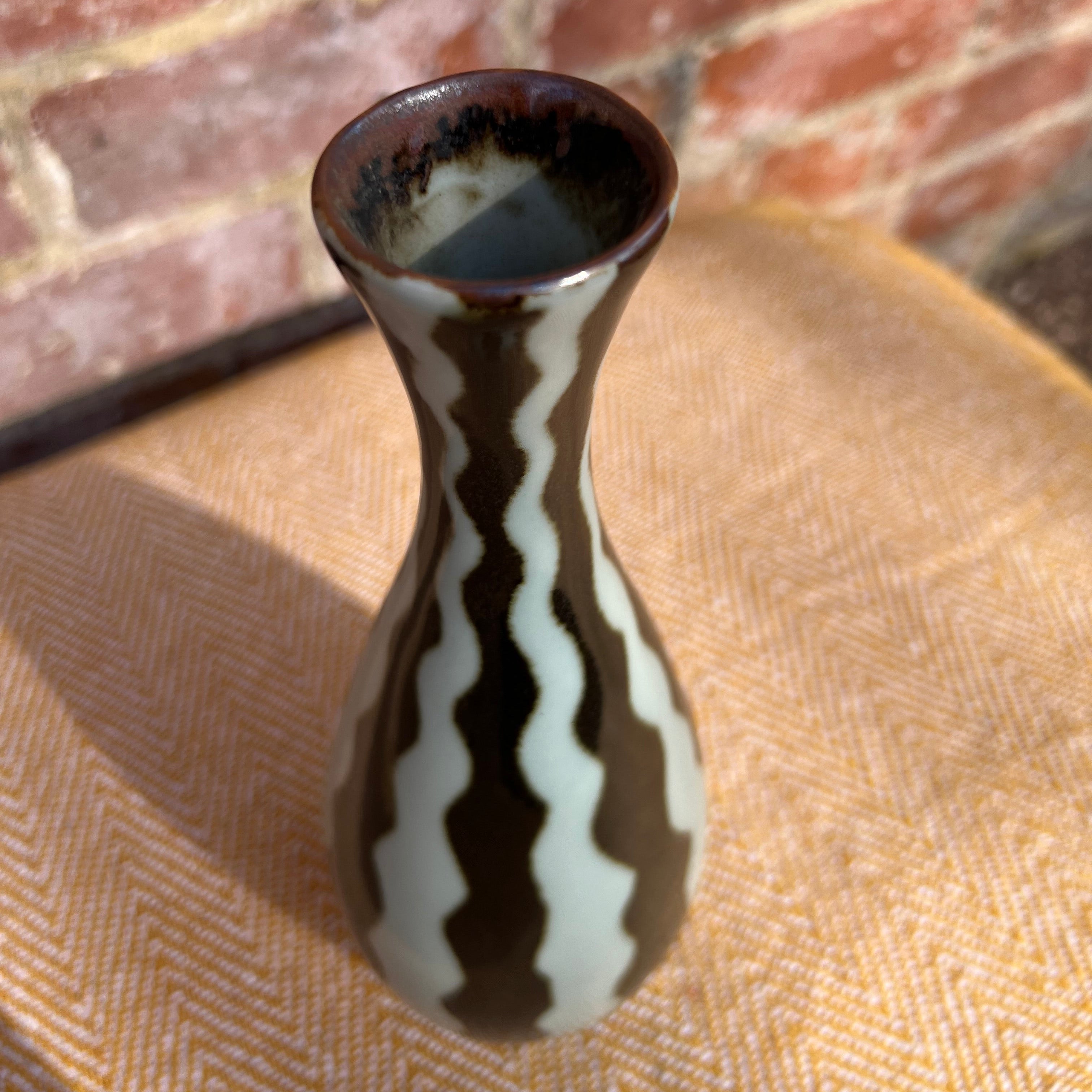 A slender studio pottery vase with white striped glossy brown and cream zigzag pattern and a flared rim