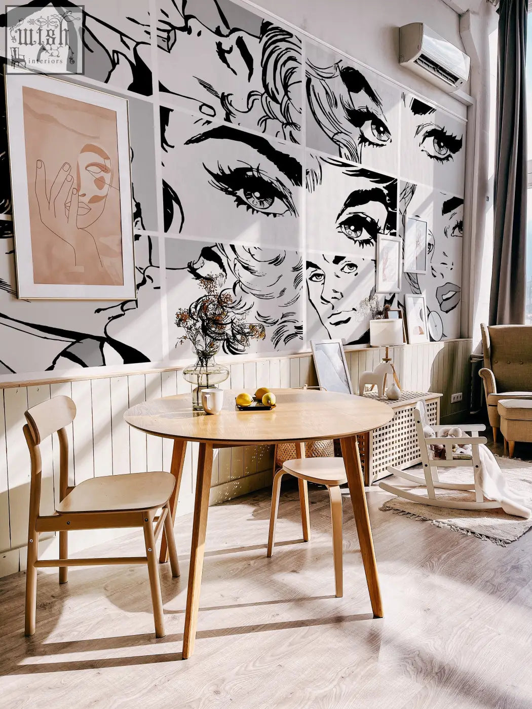 Round wooden dining table with light finish and slender legs in living space featuring black and white pop art peel and stick wallpaper