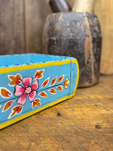 A hand-painted blue wooden brick mould with pink and orange floral designs and yellow trim used as a decorative planter or storage box