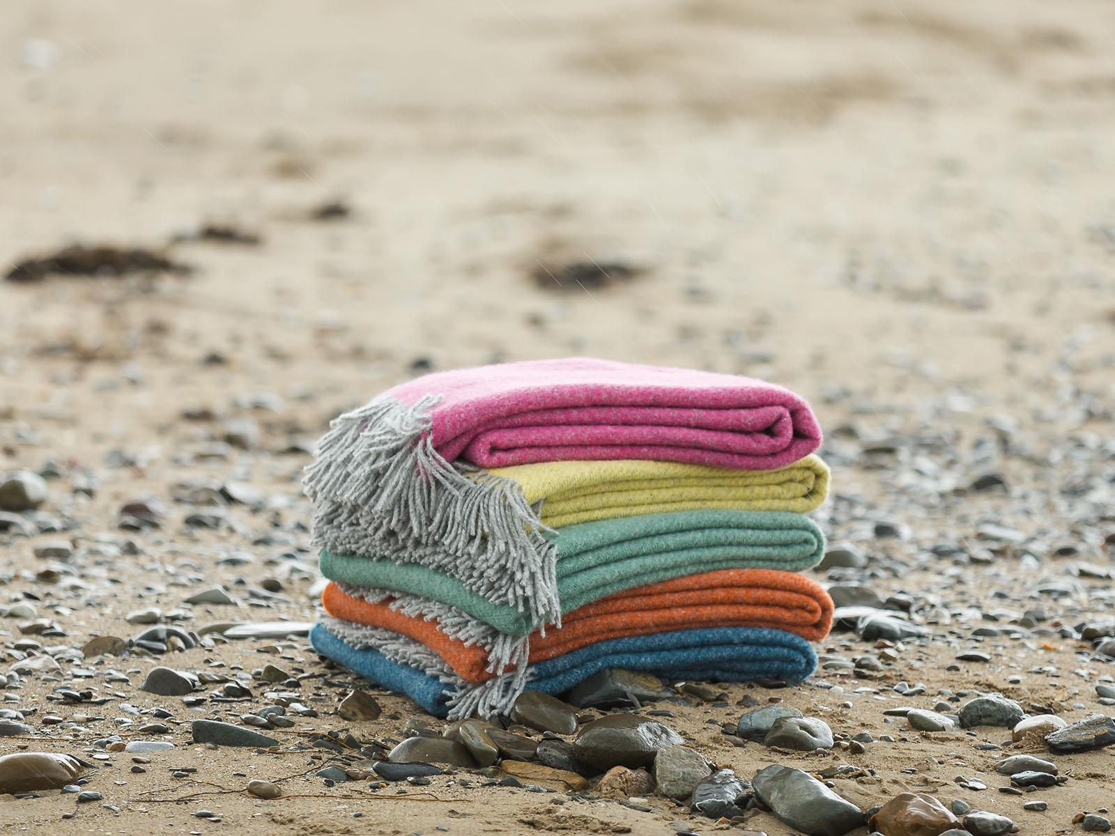A stack of five colorful boa pure new wool throw blankets in pink with fringed edges on a pebbled beach size guide