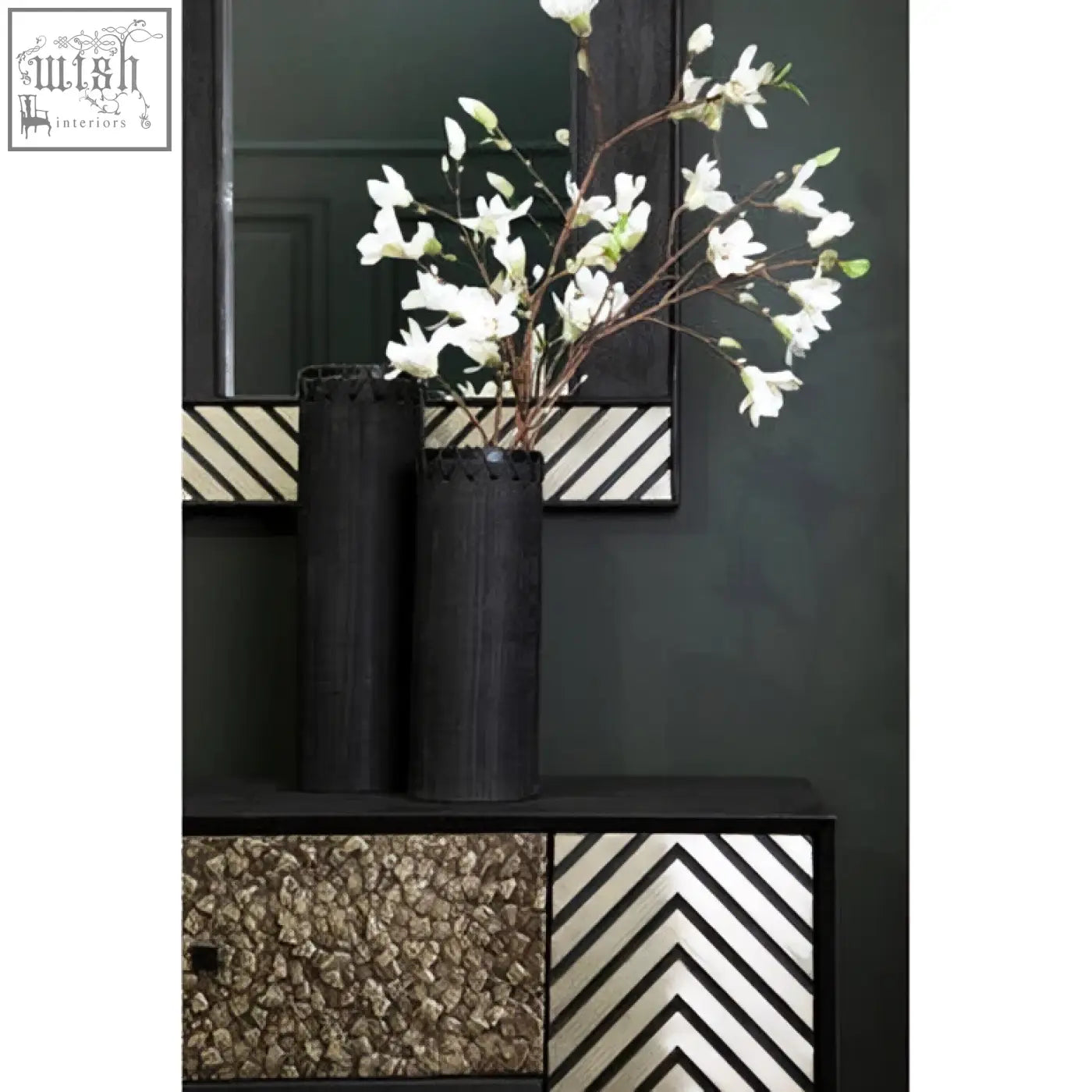 Two matte black cylindrical vases with textured rims holding white magnolia branches near a boho black wall mirror