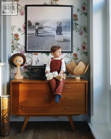 A child in rust corduroy overalls sits on a mid-century dresser in a living space with boho floral peel and stick wallpaper