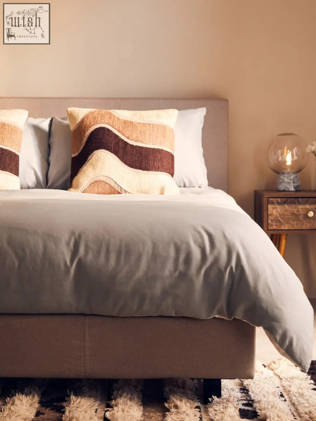 Beige upholstered bed with textured gray duvet and Bosie Ozella natural tonal cushion in earth-toned wavy stripes