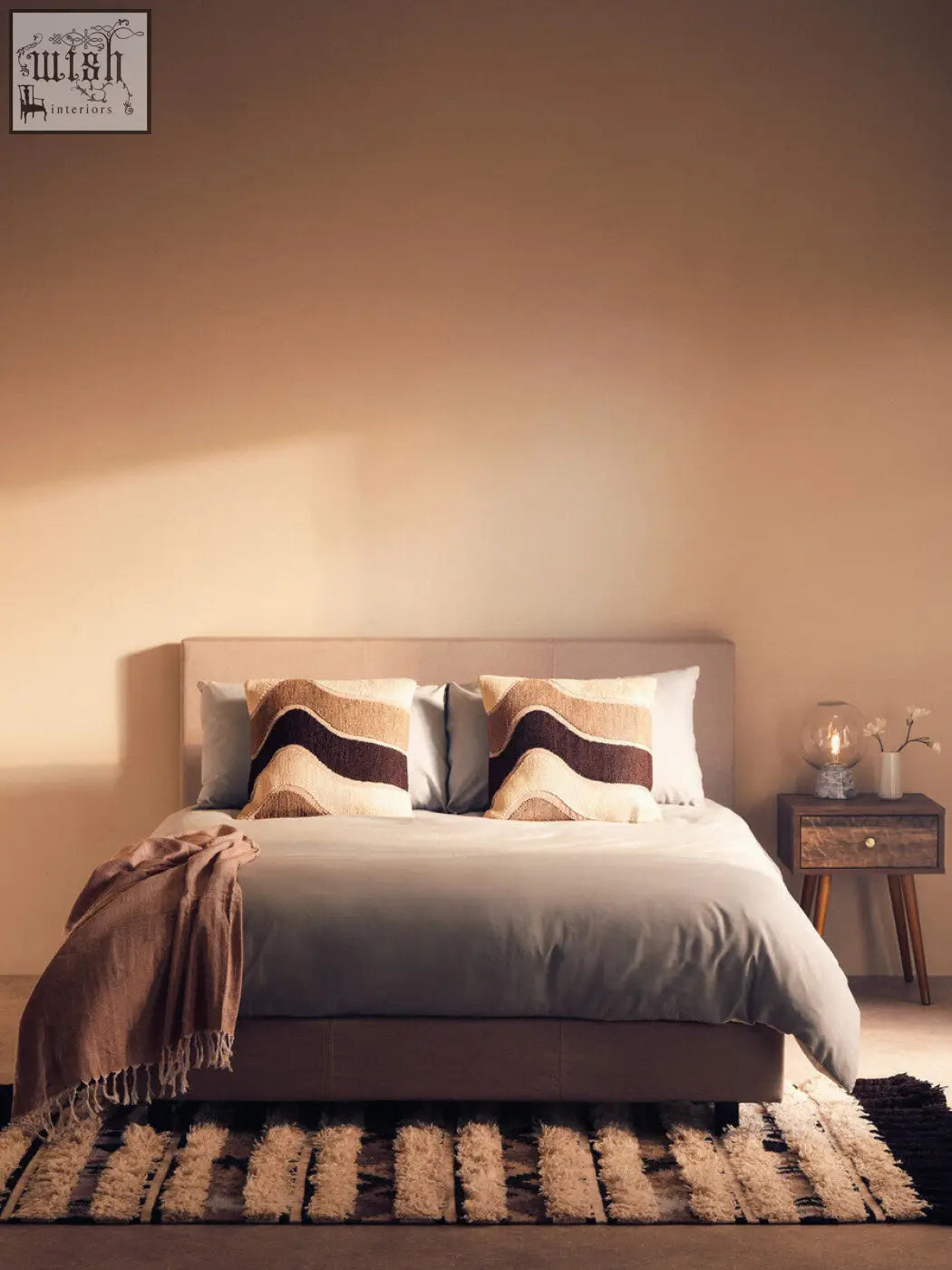 A modern upholstered bed with soft gray bedding and beige bosie ozella natural tonal cushions with brown waves