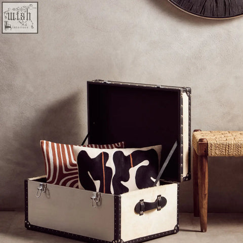 Cream-colored bosie ozella trunk with black leather trim and metal studs complements a white cushion with intricate embroidery