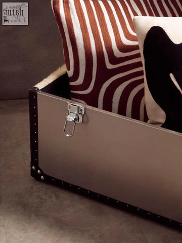 Silver metal trunk with black leather trim and rivets featuring a sturdy latch next to bosie ozella white cushion with intricate embroidery