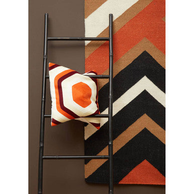 Black metal ladder with an orange white and black pillow featuring villon zig zag pattern resting on its rungs
