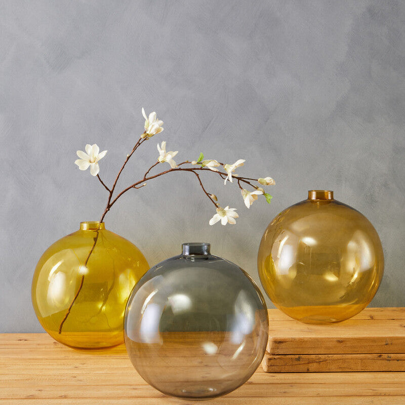 Three spherical bree grey glass vases in amber and smoky grey glass with one holding white blossom branch