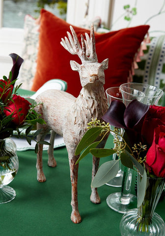 A rustic whitewashed wooden deer on green tablecloth next to a burgandy velvet cushion cover with tassels