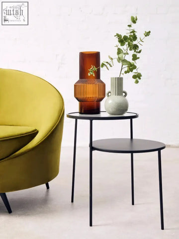 A sleek black metal side table with a round top and slender legs beside a calcita small speckled green vase