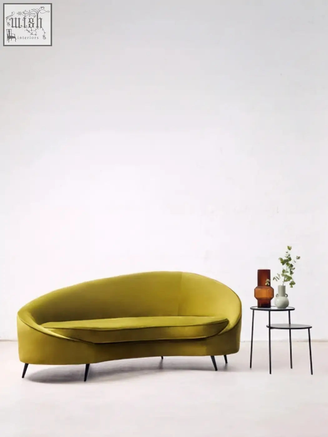 A sleek olive green velvet sofa with curved design and black metal legs next to a calcita small speckled green vase