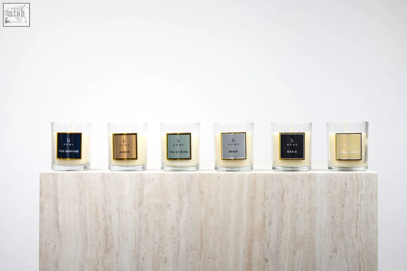 Six clear glass candle jars with colorful square labels on a marble pedestal in a candle discovery set