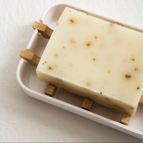 A creamy off-white bar of soap with botanical specks sits on a white ceramic soap dish with wooden block supports
