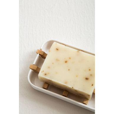 A white soap dish with four light wood clips holds an off-white white soap bar with small brown speckles