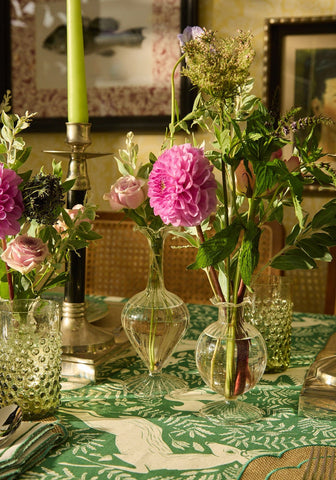 A clear glass vase with rounded body and flared base holding pink peonies next to carsons classic pair of candle holders
